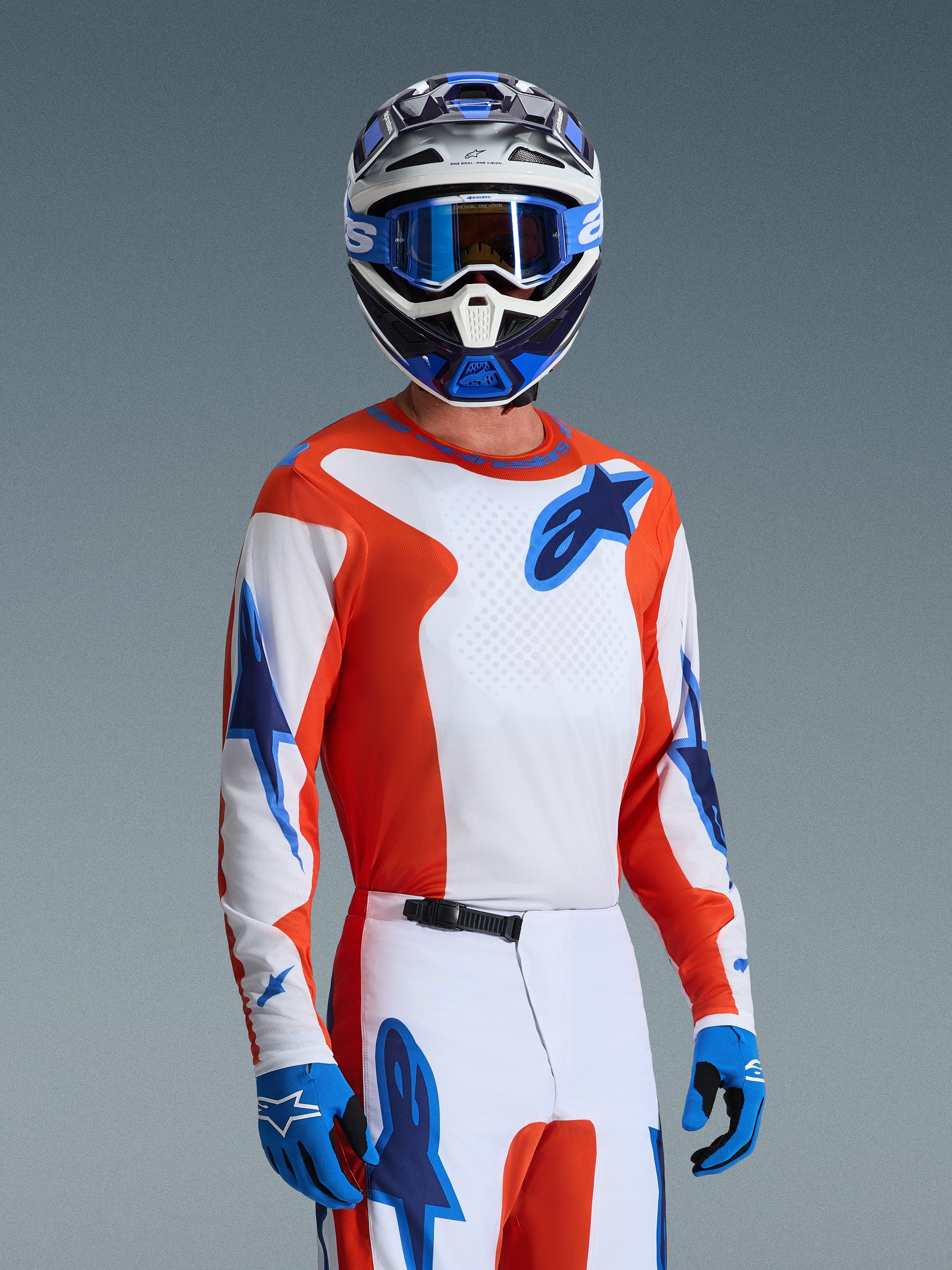 Zawodnik motocrossowy w zestawie Alpinestars 2026 Fluid Grid, pomarańczowo-biała koszulka z niebieskimi logo gwiazdy, biało-pomarańczowe spodnie z niebieskimi akcentami, niebieskie rękawice, niebiesko-biały kask z niebieskimi goglami, dopasowany sportowy strój wyścigowy do jazdy w terenie