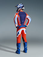 Motocyklista motocrossowy widziany od tyłu w zestawie Alpinestars 2026 Fluid Grid, pomarańczowo-niebieska koszulka z białymi rękawami, pasujące pomarańczowo-białe spodnie, niebieskie buty motocrossowe oraz niebiesko-biały kask, profesjonalny strój do wyścigów terenowych