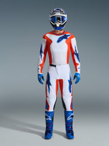 Motocross rider wearing complete racing gear, 2026 Fluid Grid Jersey in Orange and UCLA Blue, matching white pants with orange and blue accents, blue and white helmet with blue goggles, blue and purple off-road boots, blue gloves, coordinated outfit for motocross racing