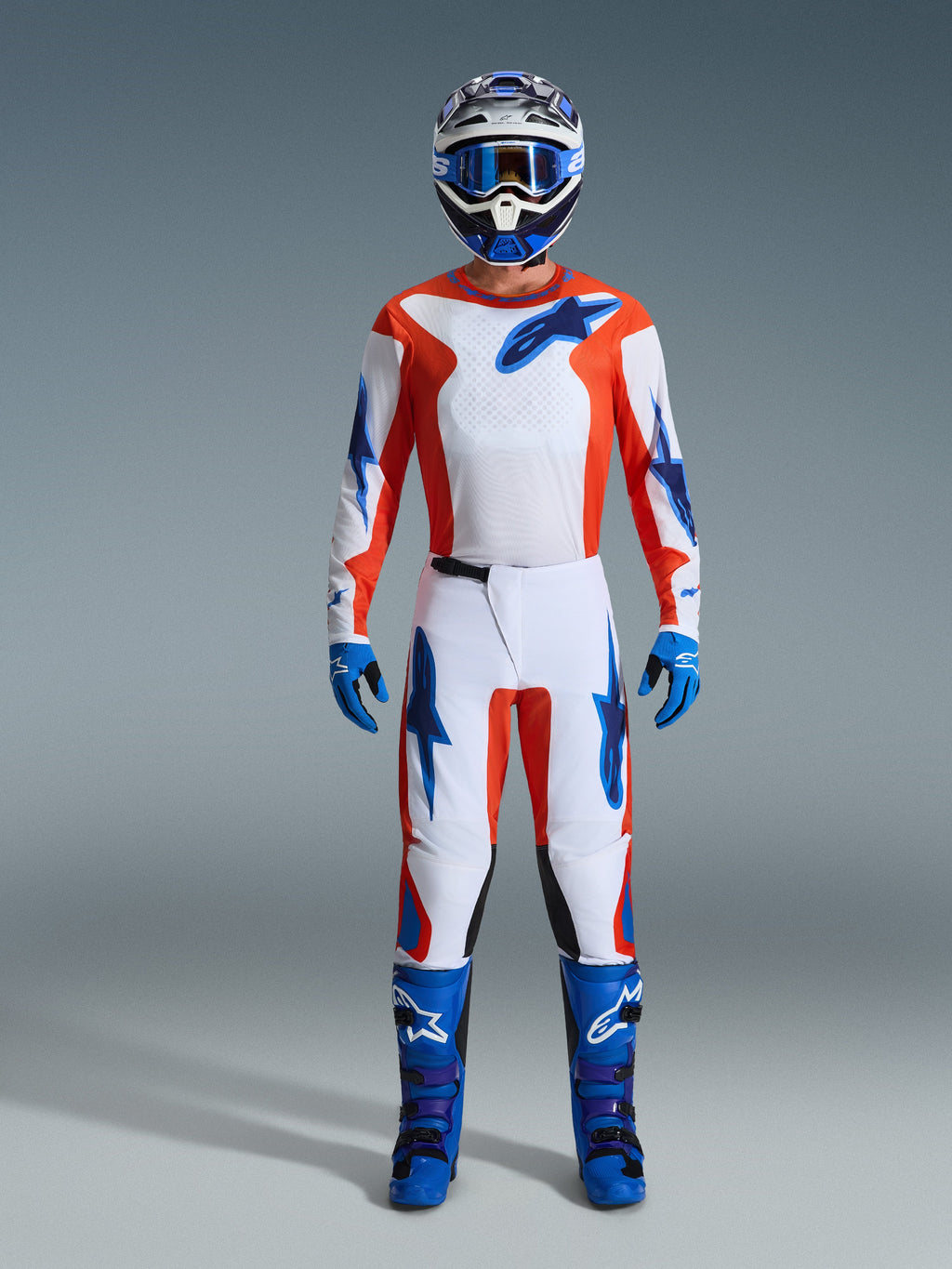 Alpinestars 2026 Fluid Grid Gear Set, motocyklista motocrossowy w pełnym rynsztunku, biała koszulka i spodnie z pomarańczowymi i błękitnymi akcentami, pasujący niebiesko-biały kask z niebieskimi goglami, niebieskie rękawice i niebieskie buty motocrossowe, dopasowany strój do wyścigów terenowych