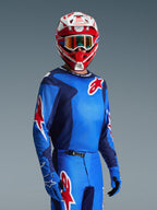 Motocyklista w koszulce Alpinestars Fluid Grid 2026, kolor niebiesko-czerwony, czerwone logo gwiazdy Alpinestars, niebieskie spodnie i rękawice, czerwono-biały kask z goglami