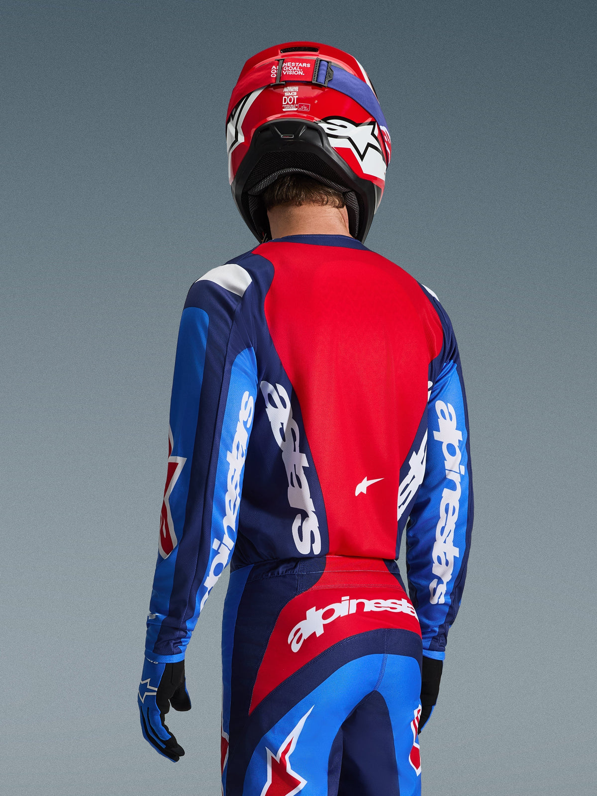 Widok z tyłu zawodnika motocrossowego w koszulce Alpinestars 2026 Fluid Grid w kolorze niebieskim i czerwonym, z wyraźnym białym logo na rękawach i plecach, w zestawieniu z pasującymi spodniami i czerwonym kaskiem, tworzący kompletny strój do wyścigów off-road
