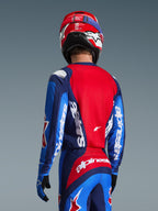 Widok z tyłu zawodnika motocrossowego w koszulce Alpinestars 2026 Fluid Grid w kolorze niebieskim i czerwonym, z wyraźnym białym logo na rękawach i plecach, w zestawieniu z pasującymi spodniami i czerwonym kaskiem, tworzący kompletny strój do wyścigów off-road