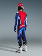 Zawodnik motocrossowy widoczny od tyłu w koszulce Alpinestars 2026 Fluid Grid w kolorze niebieskim i czerwonym, pasujące spodnie motocrossowe z wyrazistym białym brandingiem, czerwony kask oraz biało-czarne buty, kompletny zestaw odzieży wyścigowej off-road