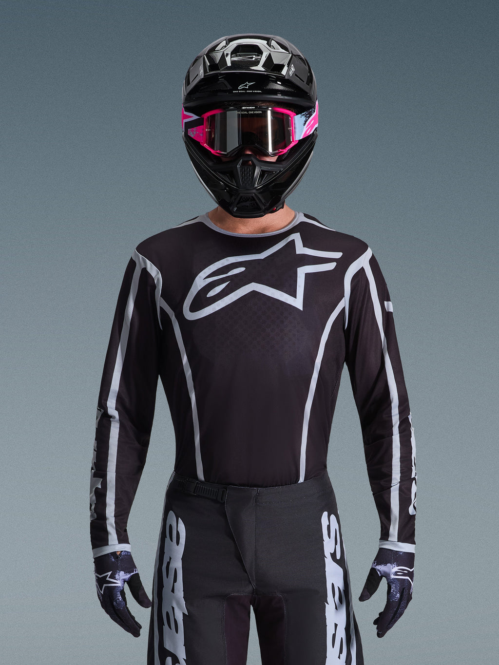 Koszulka Alpinestars 2026 Fluid Apex, koszulka motocrossowa, czarno-szara, z dużym białym logo Astars na klatce piersiowej i szarymi akcentami graficznymi na rękawach, noszona przez motocyklistę w różowych goglach i czarnym kasku, dopasowany zestaw odzieży off-road
