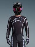 Koszulka Alpinestars 2026 Fluid Apex, koszulka motocrossowa, czarno-szara, z dużym białym logo Astars na klatce piersiowej i szarymi akcentami graficznymi na rękawach, noszona przez motocyklistę w różowych goglach i czarnym kasku, dopasowany zestaw odzieży off-road