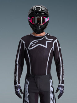 Koszulka Alpinestars 2026 Fluid Apex, koszulka motocrossowa, czarno-szara, z dużym białym logo Astars na klatce piersiowej i szarymi akcentami graficznymi na rękawach, noszona przez motocyklistę w różowych goglach i czarnym kasku, dopasowany zestaw odzieży off-road