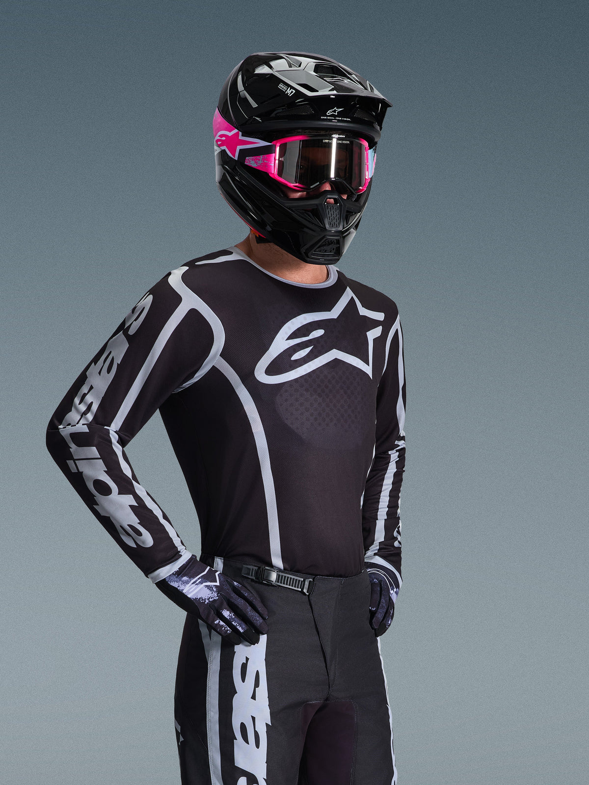 Motocyklista motocrossowy w koszulce Alpinestars 2026 Fluid Apex, czarno-szara z dużym białym logo na klatce piersiowej i geometrycznymi grafikami na rękawach, czarne spodnie, czarne rękawice, czarny kask z różowymi akcentami i różowe gogle