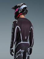 Motocrossista widoczny od tyłu w koszulce Alpinestars 2026 Fluid Apex w kolorze czarno-szarym, pasujące spodnie motocrossowe z białymi akcentami, czarny kask z różowymi logotypami, spójny strój do wyścigów off-road