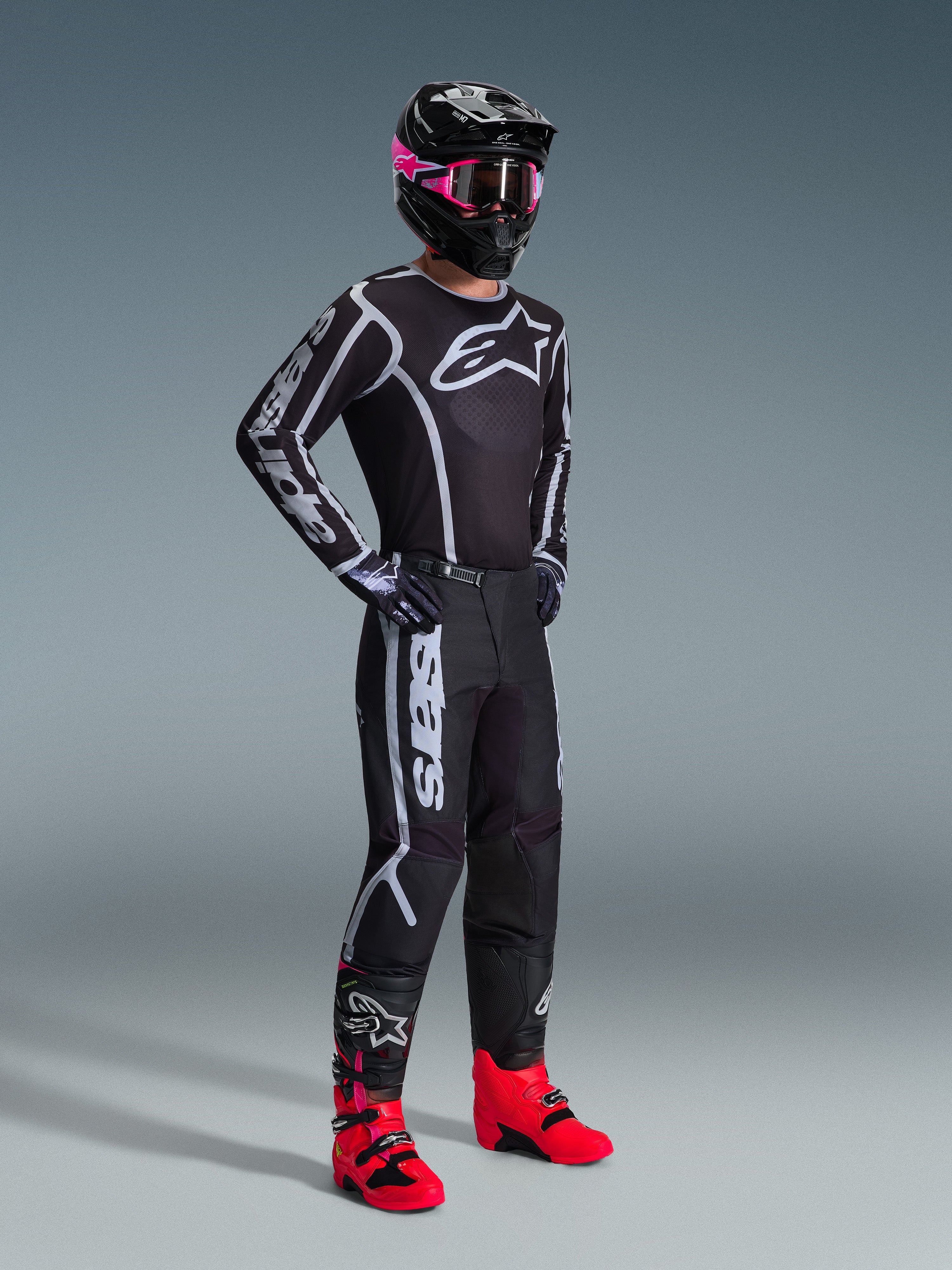 Motocyklista motocrossowy ubrany w koszulkę 2026 Fluid Apex, czarno-szara, w zestawieniu z pasującymi spodniami, czarny kask z różowymi goglami i jaskrawoczerwone buty, z wyraźnym białym brandingiem Alpinestars na klatce piersiowej i rękawach, spójny profesjonalny strój do wyścigów off-road.