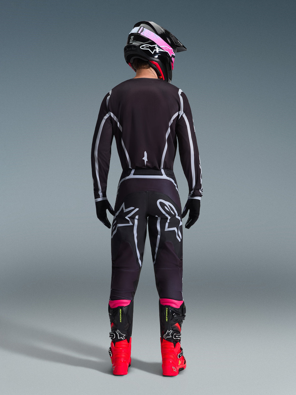 Motocyklista motocrossowy widziany od tyłu w zestawie Alpinestars 2026 Fluid Apex Gear Set, czarna bluza i spodnie z szarymi geometrycznymi liniami i logo gwiazdy, czarny kask z różowymi akcentami, jaskrawoczerwone i czarne buty, kompletny strój wyścigowy