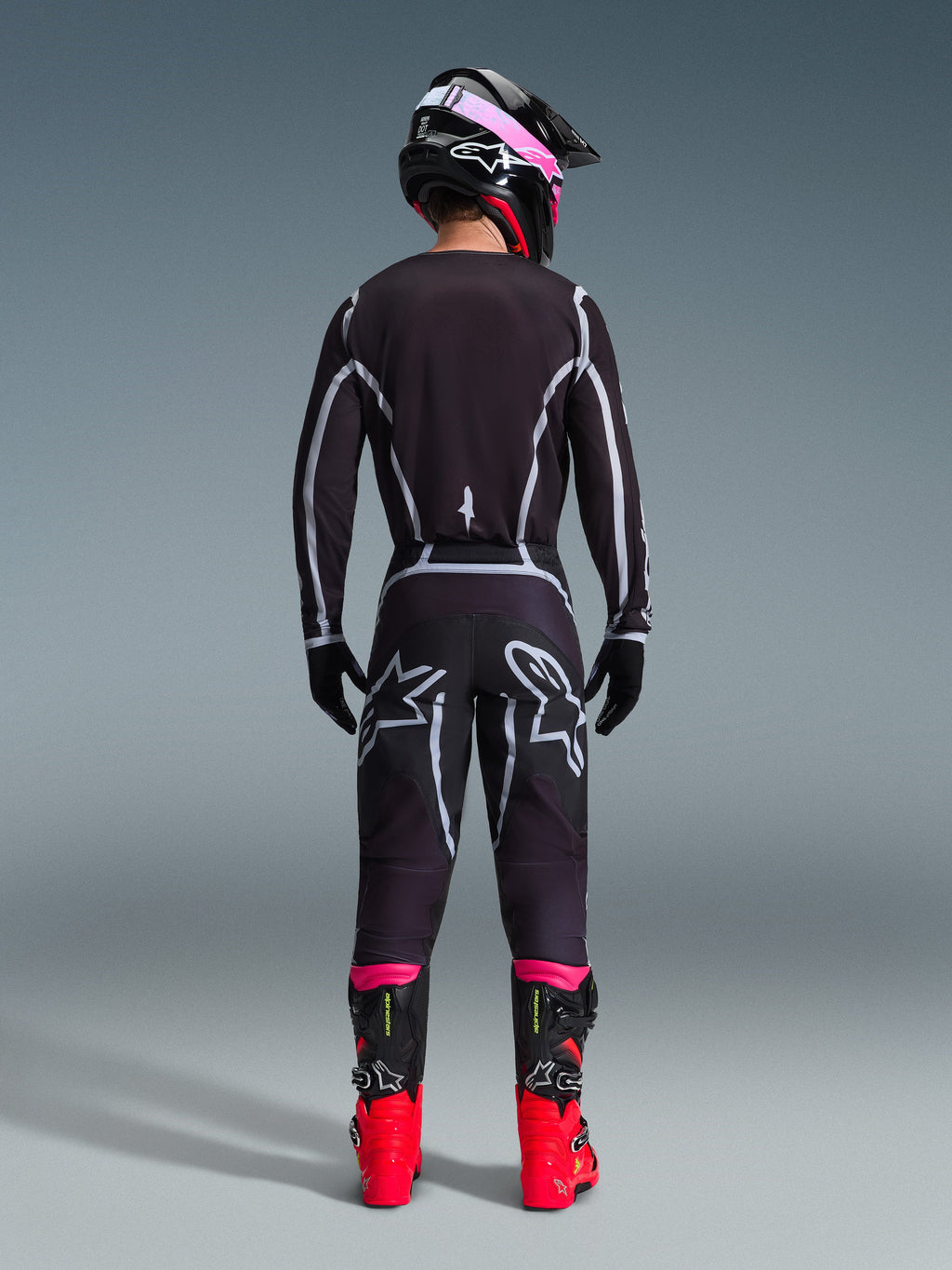 Motocyklista motocrossowy widoczny od tyłu, ubrany w koszulkę Alpinestars 2026 Fluid Apex i pasujące spodnie w kolorze czarno-szarym, z białymi geometrycznymi liniami i logo gwiazdy na siedzeniu, w czarno-różowym kasku i czerwono-czarnych butach wyścigowych.