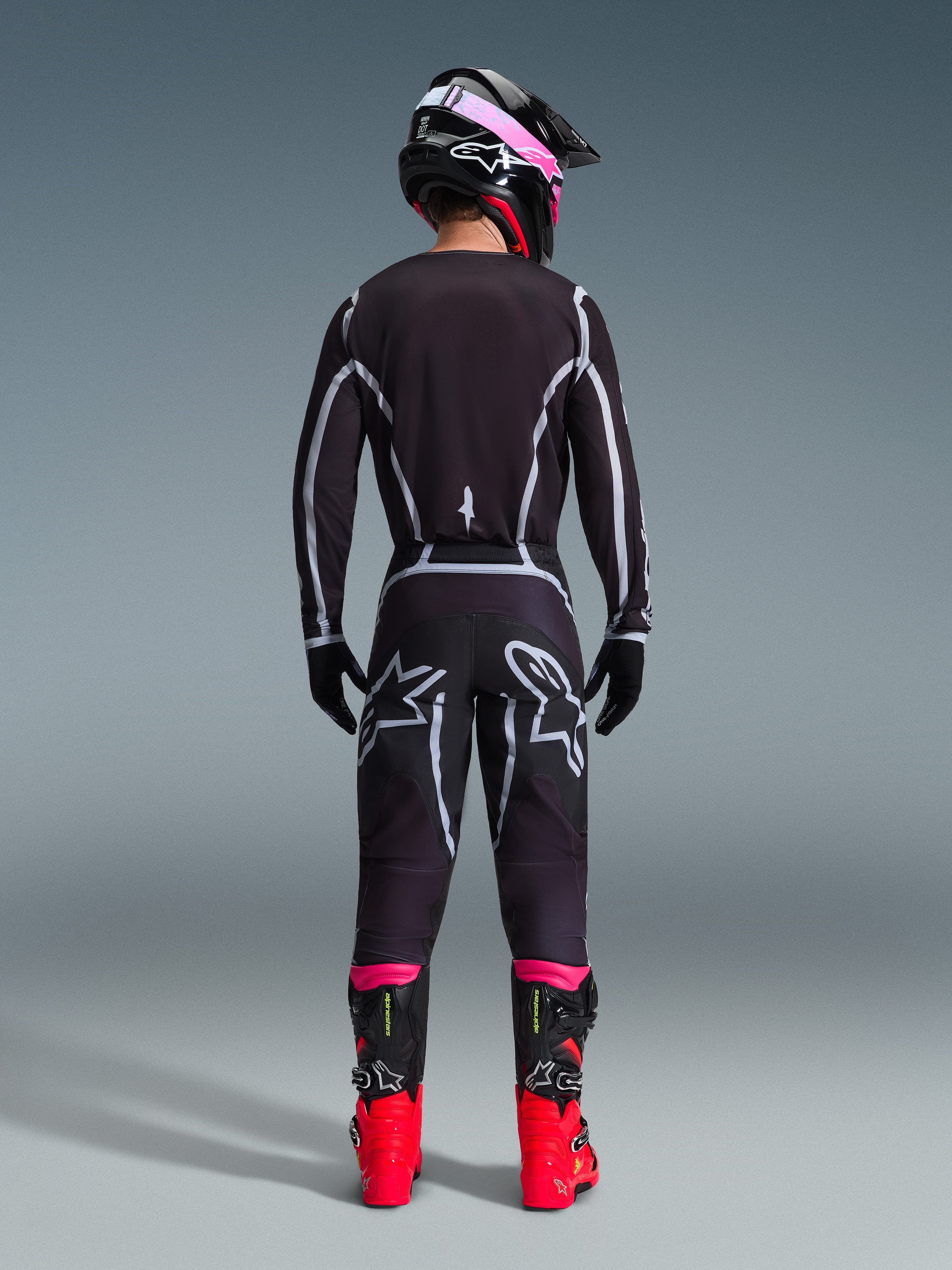 Motocyklista motocrossowy widoczny od tyłu, ubrany w koszulkę Alpinestars 2026 Fluid Apex i pasujące spodnie w kolorze czarno-szarym, z białymi geometrycznymi liniami i logo gwiazdy na siedzeniu, w czarno-różowym kasku i czerwono-czarnych butach wyścigowych.