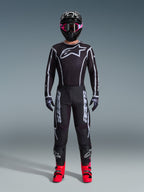 Motocyklista motocrossowy w koszulce Alpinestars 2026 Fluid Apex, czarno-szara, duże białe logo Astars na piersi, białe geometryczne linie, pasujące czarne spodnie, czarny kask z różowymi goglami, jaskraworóżowe buty, pełny strój wyścigowy motocross