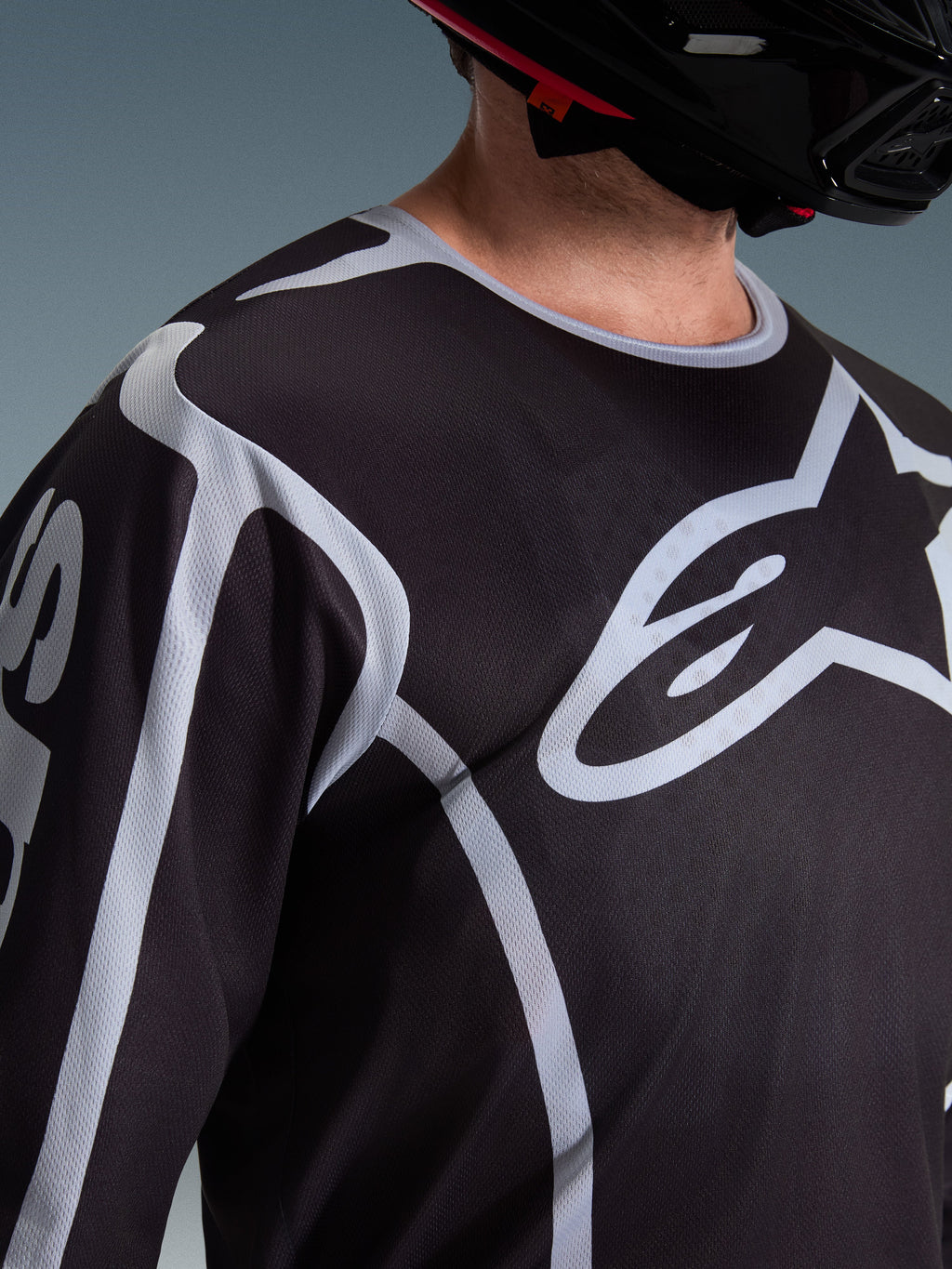 Zbliżenie na motocyklistę w koszulce Alpinestars 2026 Fluid Apex w kolorze czarno-szarym, duże białe logo Astars na klatce piersiowej, techniczna konstrukcja z siateczki, czarny kask motocrossowy