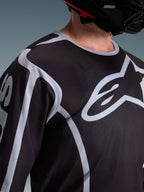 Zbliżenie na motocyklistę w koszulce Alpinestars 2026 Fluid Apex w kolorze czarno-szarym, duże białe logo Astars na klatce piersiowej, techniczna konstrukcja z siateczki, czarny kask motocrossowy