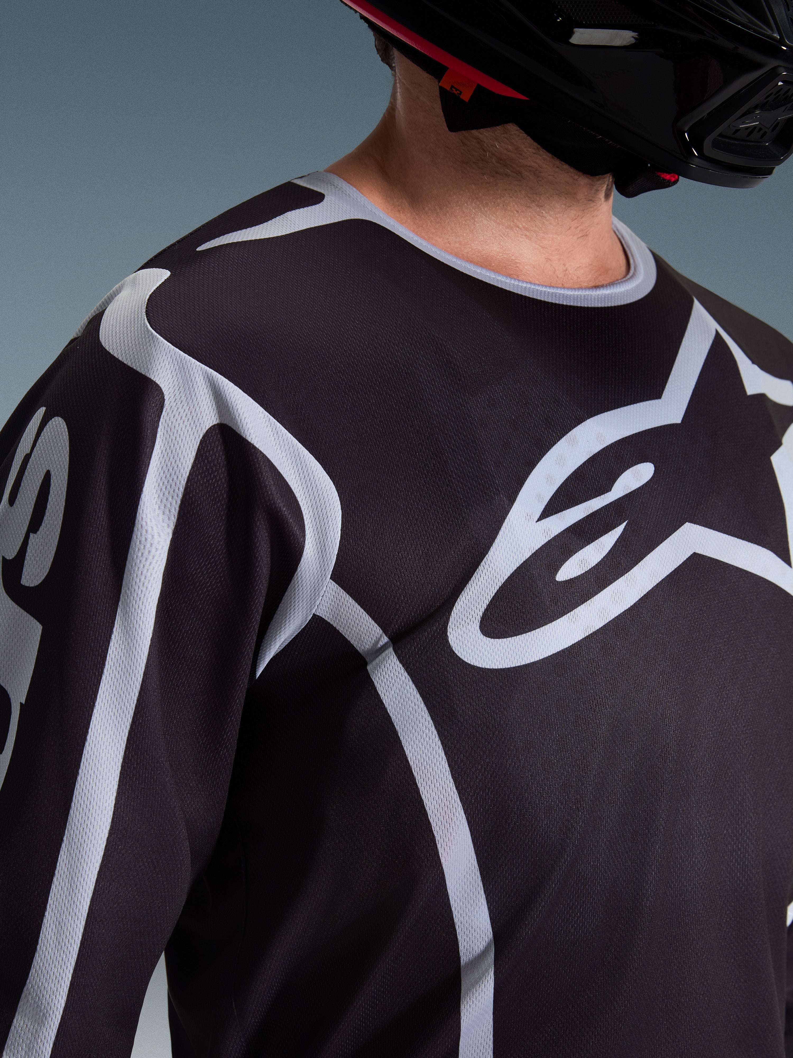 Zbliżenie na motocyklistę w koszulce Alpinestars 2026 Fluid Apex w kolorze czarno-szarym, duże białe logo Astars na klatce piersiowej, techniczna konstrukcja z siateczki, czarny kask motocrossowy