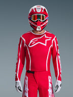 Zawodnik motocrossowy w kompletnym zestawie Alpinestars 2026 Fluid Apex, czerwona koszulka z dużym białym logo gwiazdy na piersi, pasujące czerwone spodnie z białym logowaniem, czerwono-biały kask z opalizującymi goglami, biało-czarne rękawice, dopasowany czerwono-biały strój do wyścigów terenowych