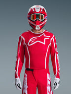 Alpinestars 2026 Fluid Apex Jersey, koszulka motocrossowa, czerwono-biała, noszona przez motocyklistę w dopasowanym czerwono-białym kasku i pomarańczowych goglach, z wyraźnym białym logo na piersi, sportowy krój, element spójnego zestawu odzieży wyścigowej off-road