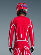 Widok z tyłu zawodnika motocrossowego w koszulce Alpinestars 2026 Fluid Apex w kolorze czerwono-białym, z białymi geometrycznymi wzorami, zestawionej z dopasowanymi spodniami motocrossowymi i czerwonym kaskiem; kompletny strój wyścigowy do jazdy terenowej.