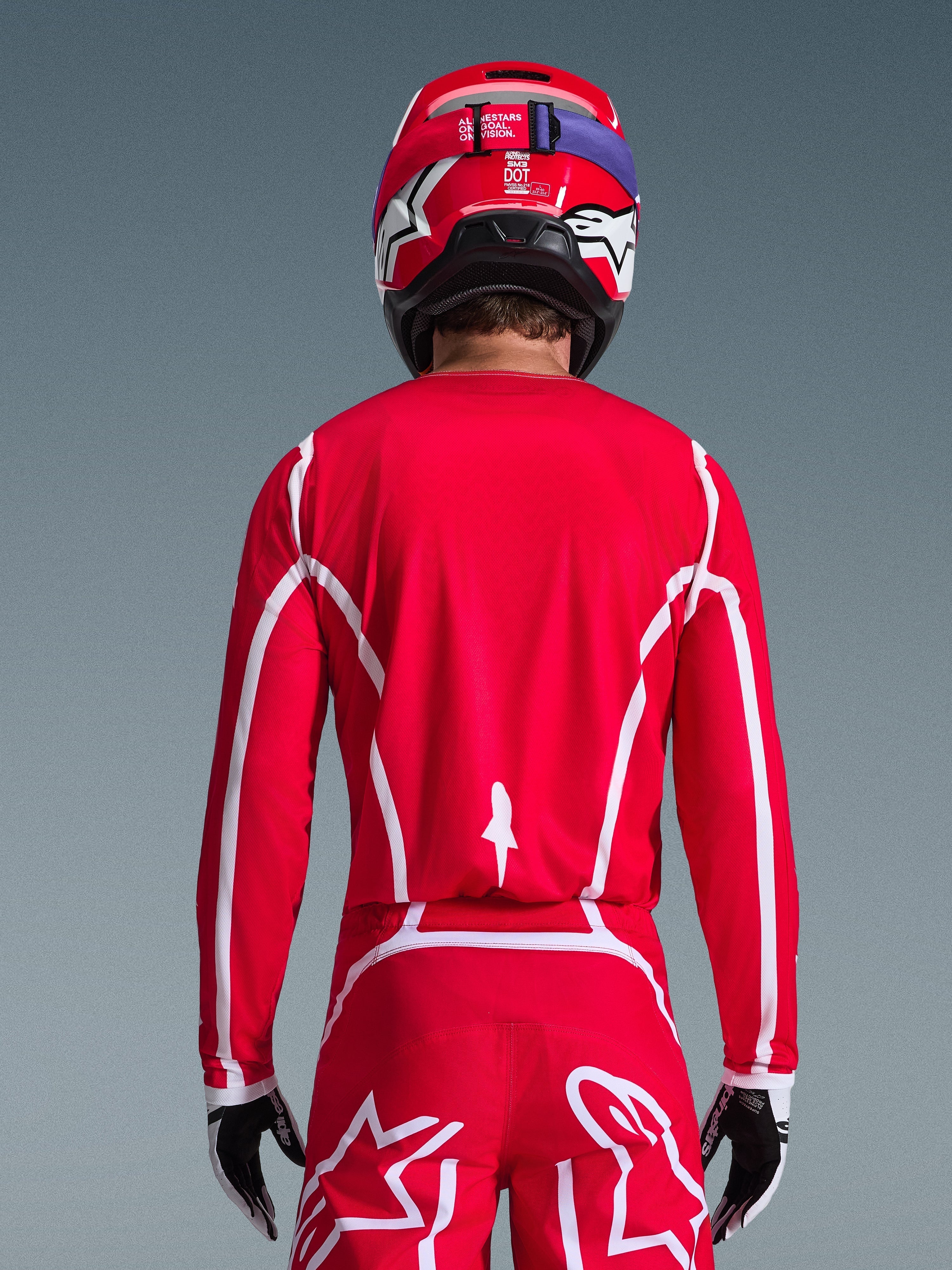 Widok z tyłu zawodnika motocrossowego w koszulce Alpinestars 2026 Fluid Apex w kolorze czerwono-białym, z białymi geometrycznymi wzorami, zestawionej z dopasowanymi spodniami motocrossowymi i czerwonym kaskiem; kompletny strój wyścigowy do jazdy terenowej.