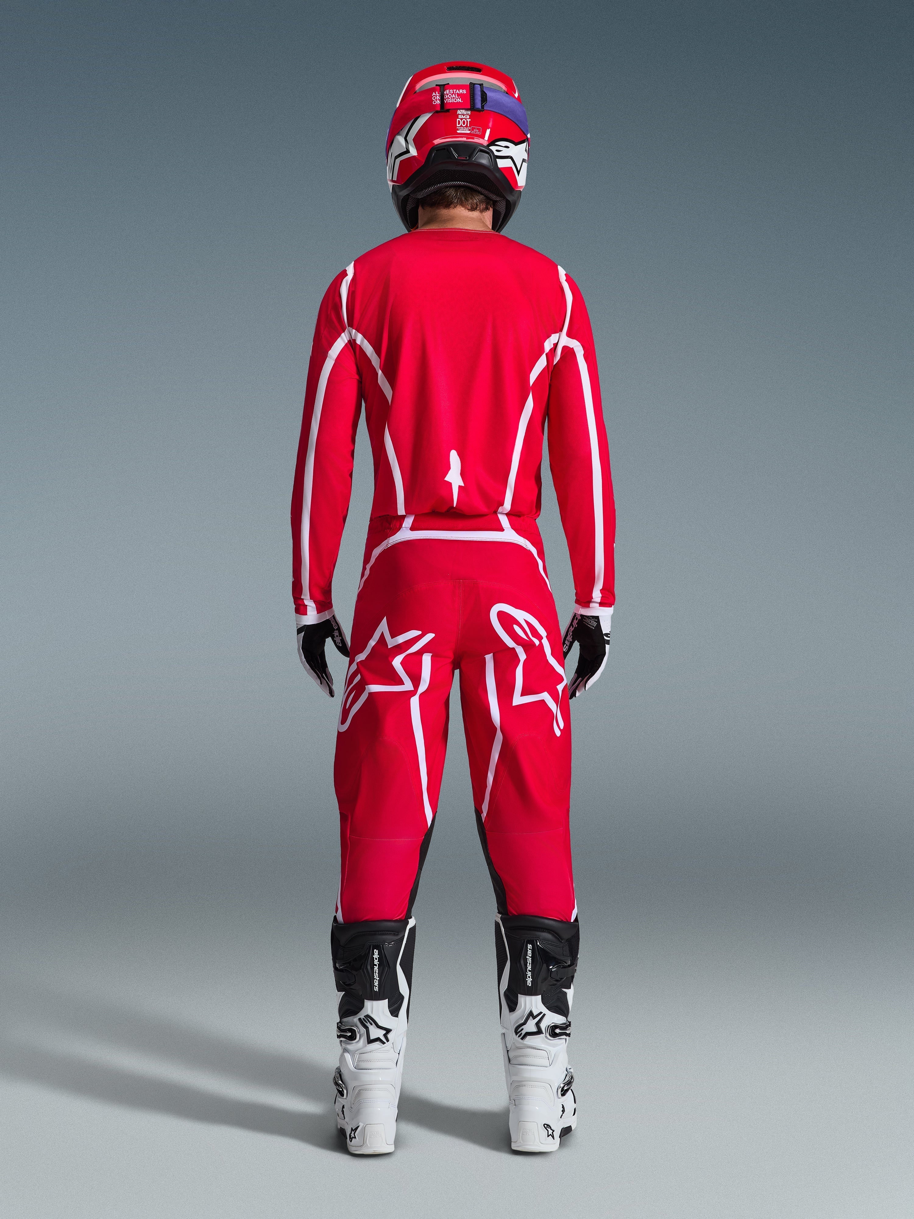 Motocyklista motocrossowy widziany od tyłu w koszulce Alpinestars 2026 Fluid Apex w kolorze czerwono-białym, dopasowane czerwone spodnie z wyraźnym białym logo gwiazdy, czerwony kask, biało-czarne buty motocrossowe i czarno-białe rękawice, kompletny zestaw do wyścigów off-road