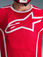 Zbliżenie na koszulkę Alpinestars 2026 Fluid Apex w kolorze czerwono-białym, z dużym białym logo Astars na klatce piersiowej, oddychającą konstrukcją z siatki poliestrowej i białym wykończeniem kołnierzyka, odzież motocrossowa