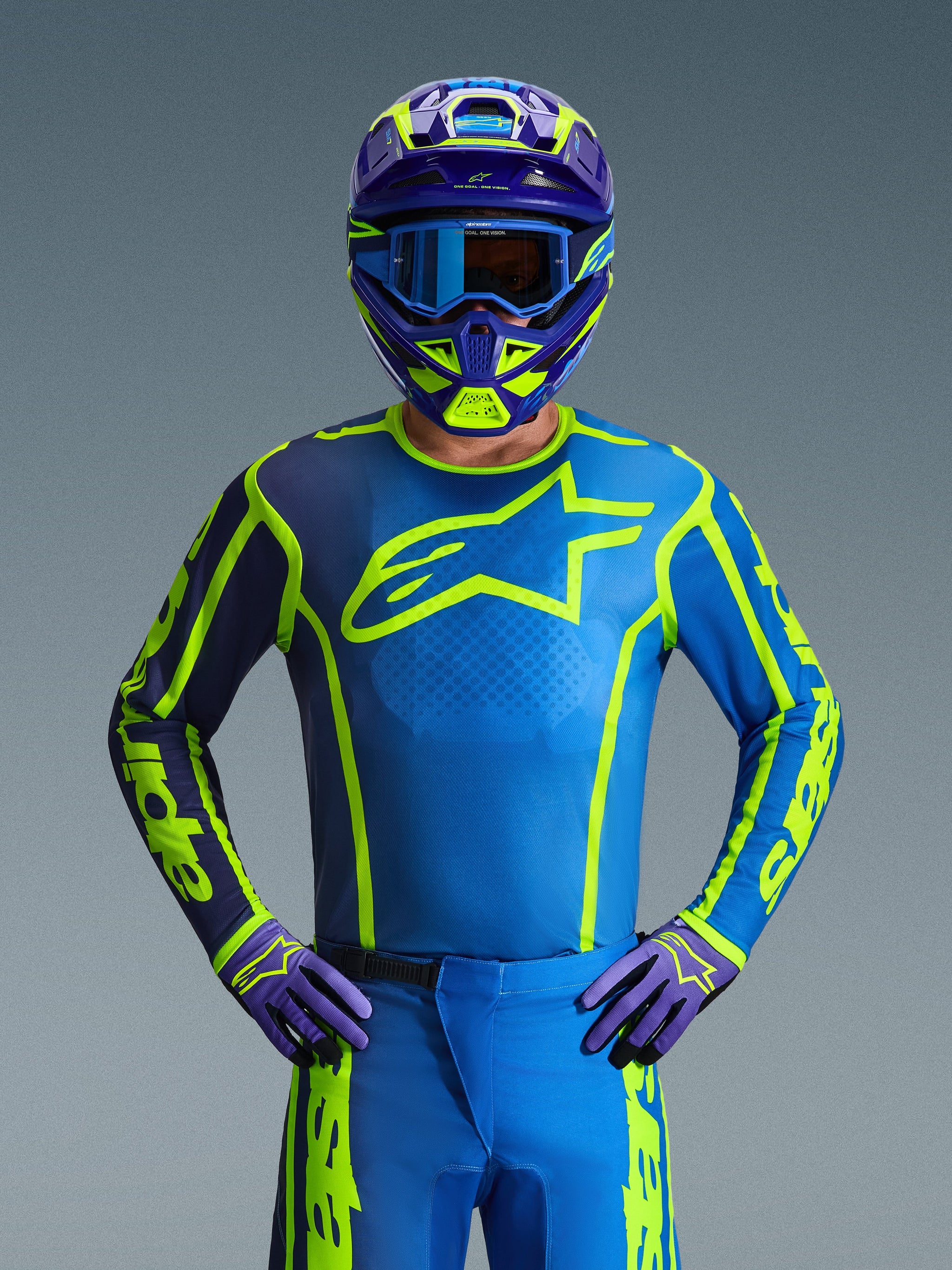 Zawodnik motocrossowy w koszulce Alpinestars 2026 Fluid Apex w kolorze Blue Yellow Fluo, z intensywnie niebieską bazą, jaskrawożółtymi grafikami i dużym logo na piersi, w dopasowanym kasku i rękawicach, zaprojektowanej do jazdy off-road i wyścigów motocrossowych