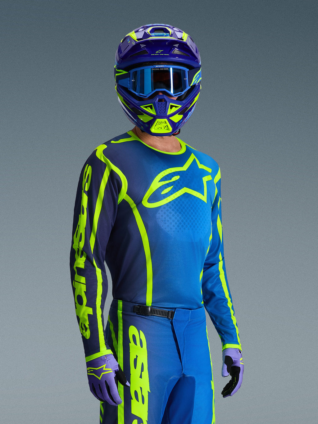 Motocyklista motocrossowy w koszulce Alpinestars Fluid Apex 2026 w kolorze niebieskim i żółtym fluo, pasujące niebieskie i neonowe żółte spodnie, fioletowo-żółty kask z niebieskimi goglami i dopasowane rękawice, kompletny strój wyścigowy off-road.