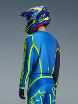 Alpinestars 2026 Fluid Apex Jersey, niebiesko-żółty fluo, widok od tyłu zawodnika w dopasowanych spodniach motocrossowych i kasku, fluorescencyjne żółte grafiki geometryczne, swobodny sportowy krój, skoordynowany zestaw odzieży do wyścigów terenowych