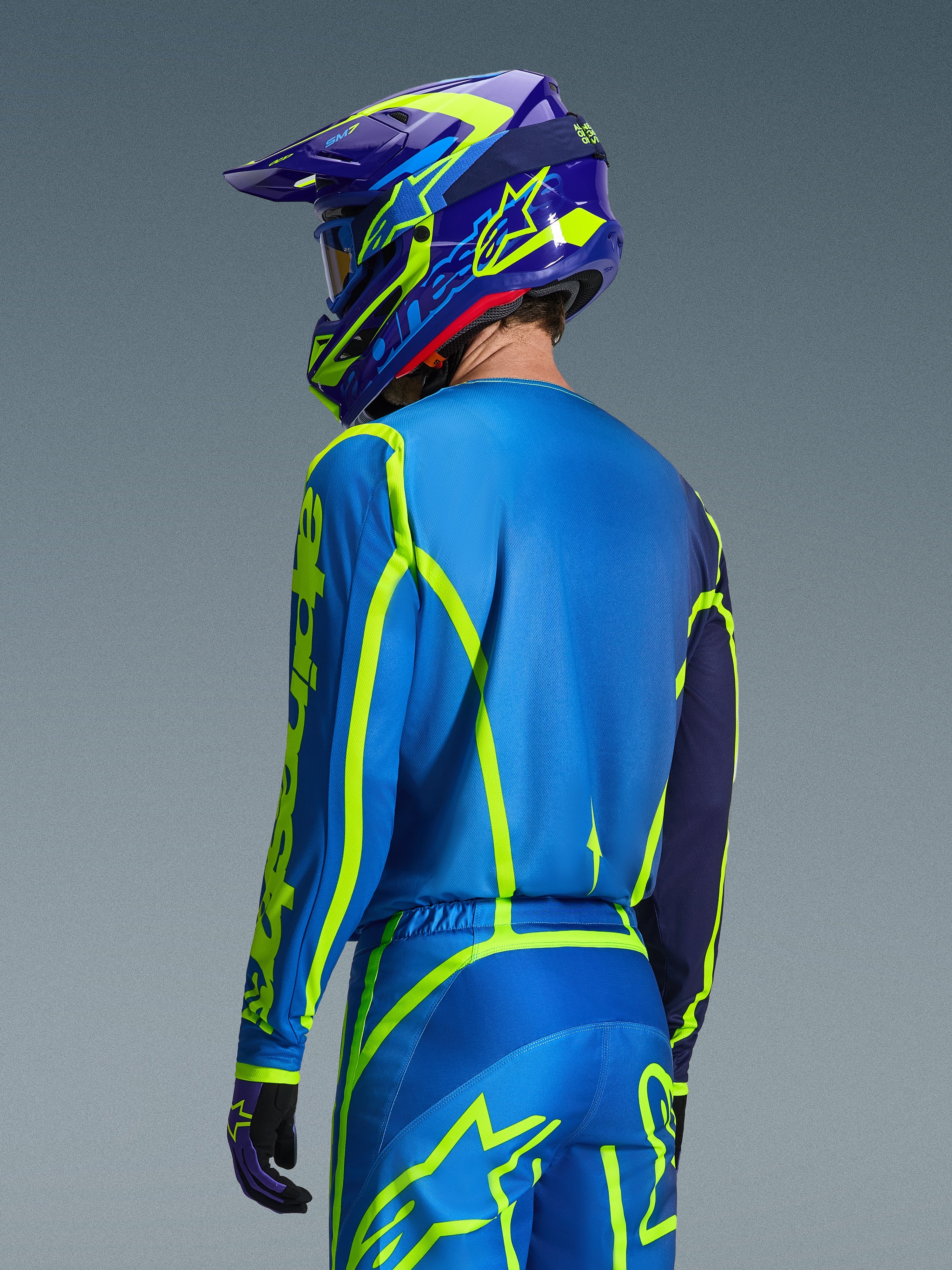 Alpinestars 2026 Fluid Apex Jersey, niebiesko-żółty fluo, widok od tyłu zawodnika w dopasowanych spodniach motocrossowych i kasku, fluorescencyjne żółte grafiki geometryczne, swobodny sportowy krój, skoordynowany zestaw odzieży do wyścigów terenowych