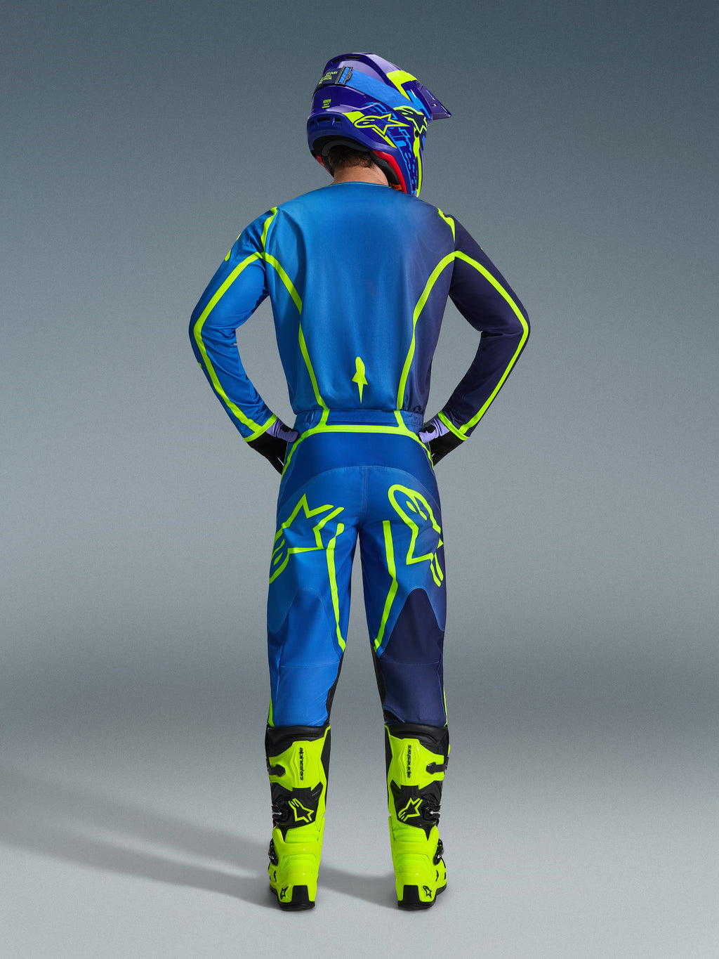 Zestaw Alpinestars 2026 Fluid Apex, widok od tyłu zawodnika motocrossowego w niebiesko-granatowej koszulce i pasujących spodniach z jaskrawymi neonowo-zielonymi akcentami, neonowo-żółte buty i fioletowy kask, profesjonalny strój wyścigowy off-road.