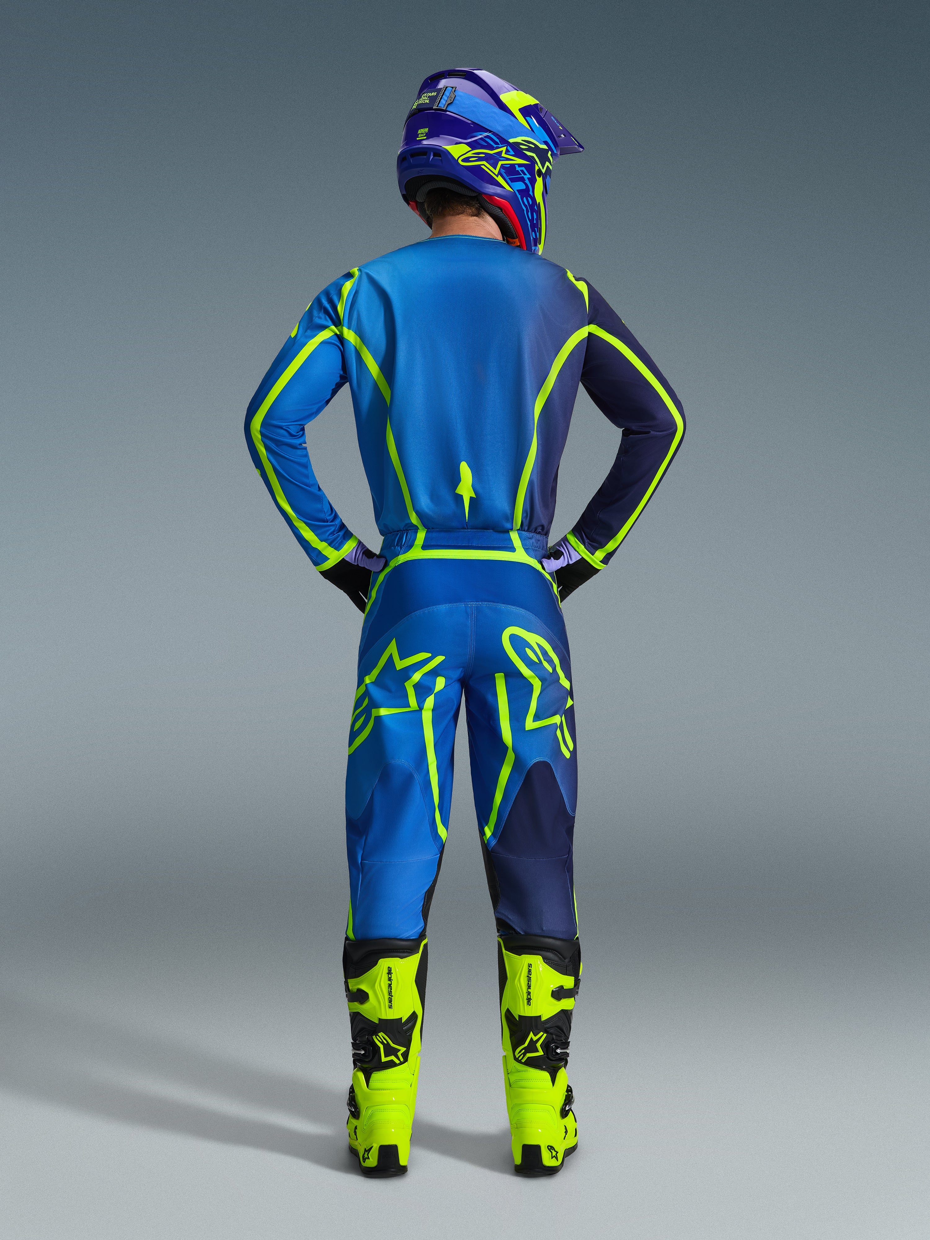 Motocyklista motocrossowy widziany od tyłu w pełnym stroju wyścigowym, koszulka Alpinestars 2026 Fluid Apex i dopasowane spodnie w kolorze Blue Yellow Fluo, neonowe akcenty i wyraźne logo marki, jasnożółte buty i dopasowany kask