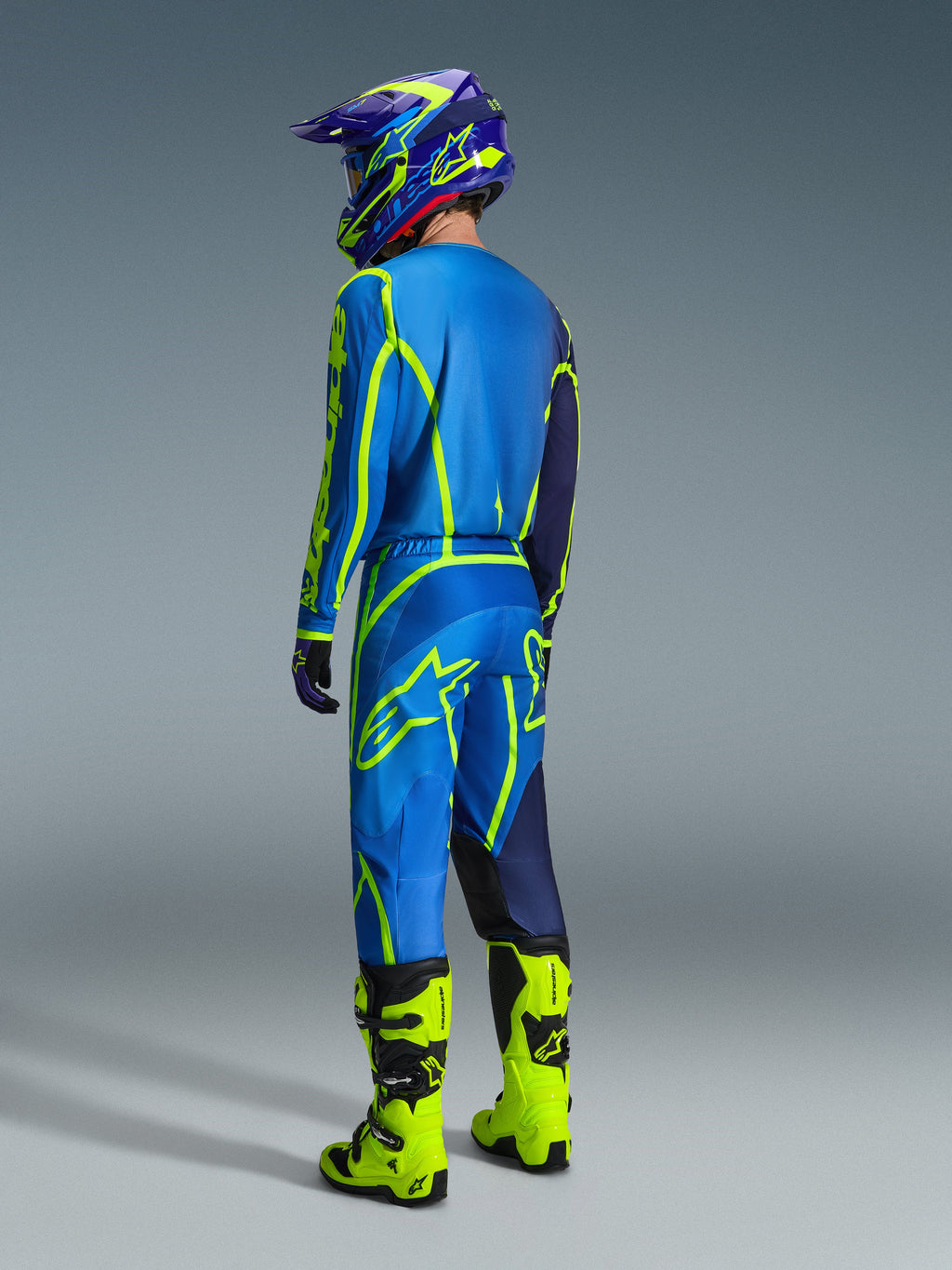 Kierowca motocrossowy widziany od tyłu, ubrany w koszulkę Alpinestars 2026 Fluid Apex w kolorze niebiesko-żółtym fluo, pasujące niebieskie i fluorescencyjne żółte spodnie, jaskrawożółte buty oraz dopasowany kask, profesjonalny sprzęt do wyścigów off-road