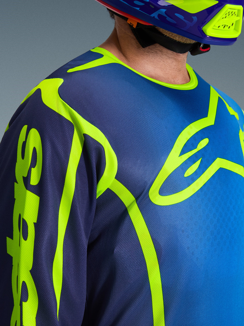 Zbliżenie na koszulkę Alpinestars 2026 Fluid Apex w kolorze niebieskim i żółtym fluo, z dużym neonowożółtym logo Astars na piersi, pasującym kaskiem motocrossowym i konstrukcją z odprowadzającego wilgoć materiału poliestrowego.
