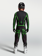 Koszulka Alpinestars Fluid Apex V2, czarno-zielona, widok z tyłu na motocyklistę w białych butach, odzież motocrossowa.