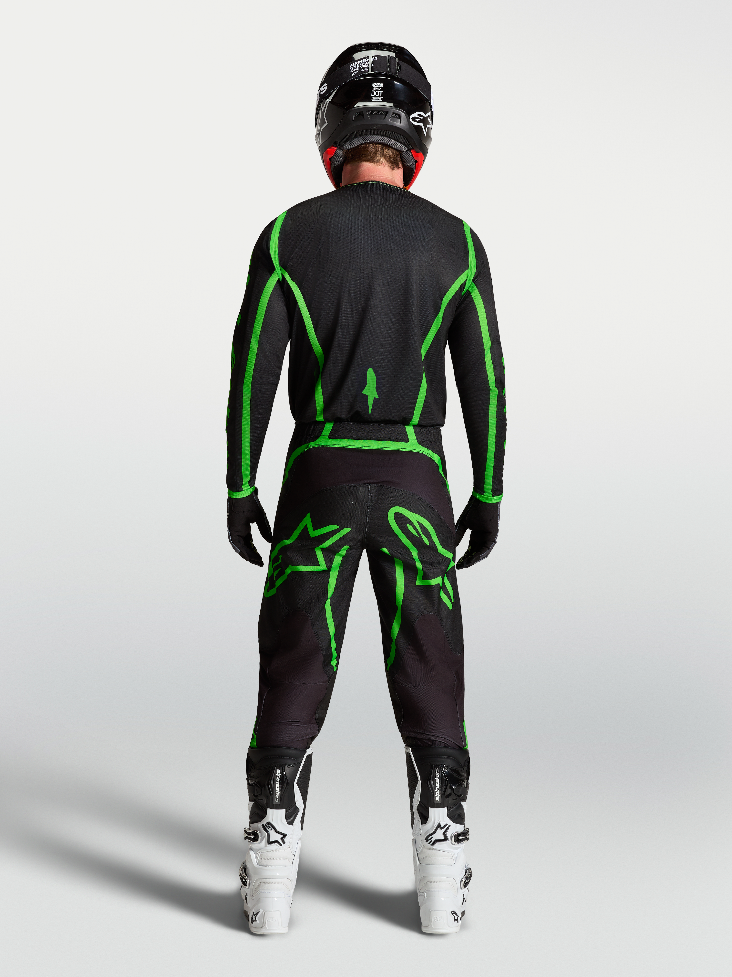 Koszulka Alpinestars Fluid Apex V2, czarno-zielona, widok z tyłu na motocyklistę w białych butach, odzież motocrossowa.