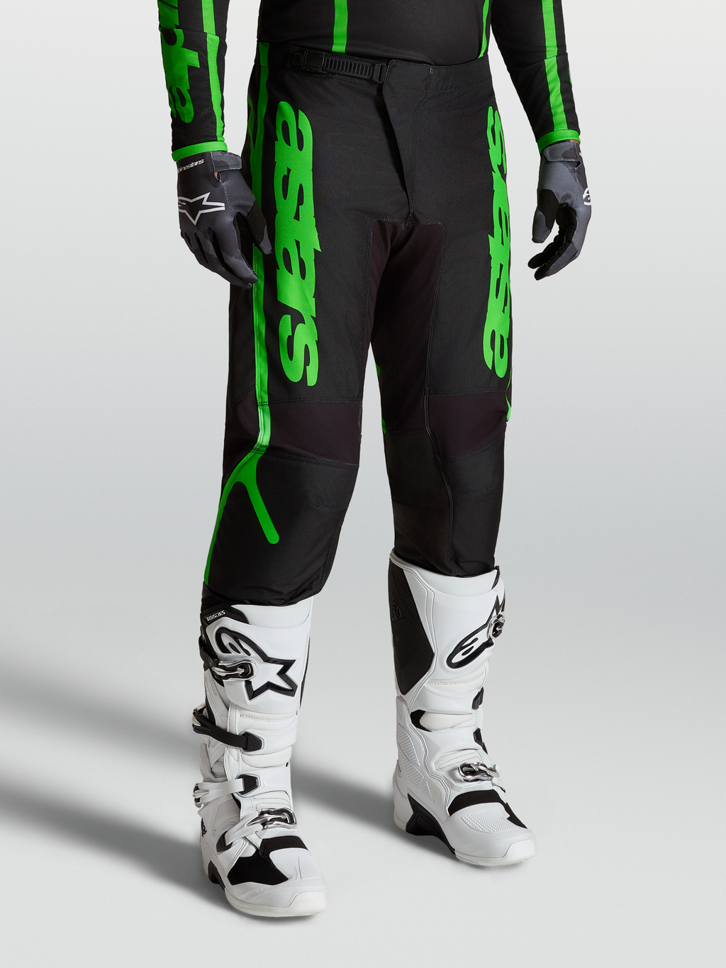 Spodnie Alpinestars Fluid Apex V2, męskie spodnie motocrossowe, czarne i fluorescencyjne zielone, widok na model z białymi butami Tech 7 i rękawicami, wyczynowy sprzęt off-road z wzmocnionego poliestru 600D.