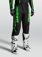 Spodnie Alpinestars Fluid Apex V2, męskie spodnie motocrossowe, czarne i fluorescencyjne zielone, widok na model z białymi butami Tech 7 i rękawicami, wyczynowy sprzęt off-road z wzmocnionego poliestru 600D.