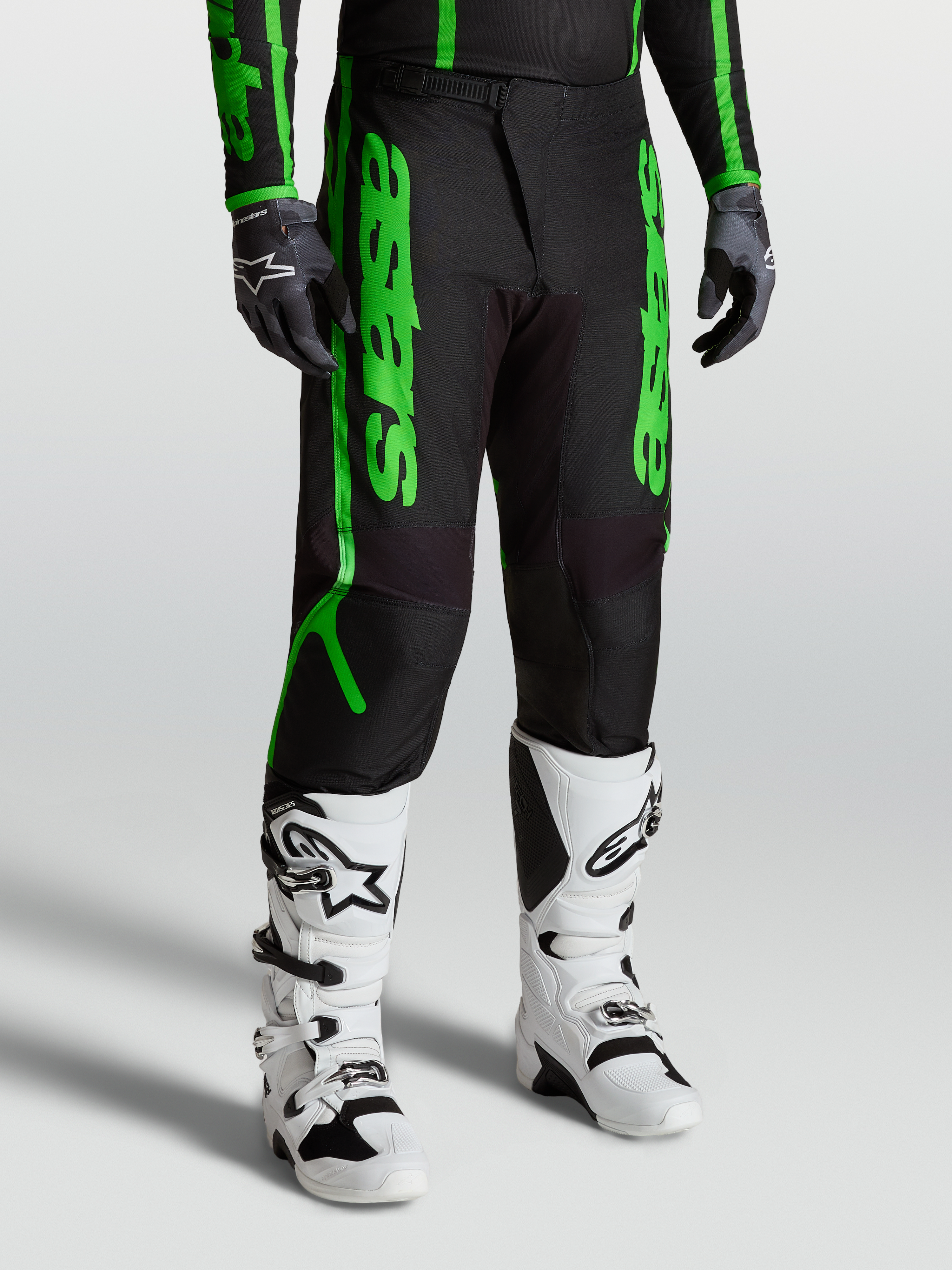 Spodnie Alpinestars Fluid Apex V2, męskie spodnie motocrossowe, czarne i fluorescencyjne zielone, widok na model z białymi butami Tech 7 i rękawicami, wyczynowy sprzęt off-road z wzmocnionego poliestru 600D.