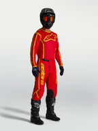 Alpinestars Koszulka Fluid Apex V2, męska koszulka motocrossowa, czerwona i złota, lekki materiał odprowadzający wilgoć, sportowy krój off-road, model w pełnym wyposażeniu ochronnym z kaskiem i butami.