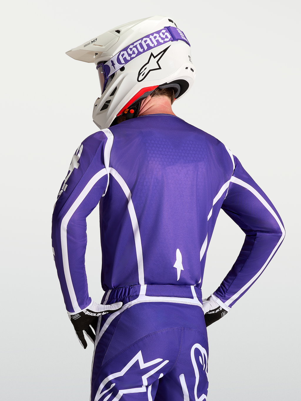 Alpinestars Koszulka Fluid Apex V2, męska koszulka motocrossowa, fioletowa i biała, oddychająca konstrukcja z poliestru, widok od tyłu na zawodnika w kasku i rękawicach.