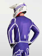 Alpinestars Koszulka Fluid Apex V2, męska koszulka motocrossowa, fioletowa i biała, oddychająca konstrukcja z poliestru, widok od tyłu na zawodnika w kasku i rękawicach.