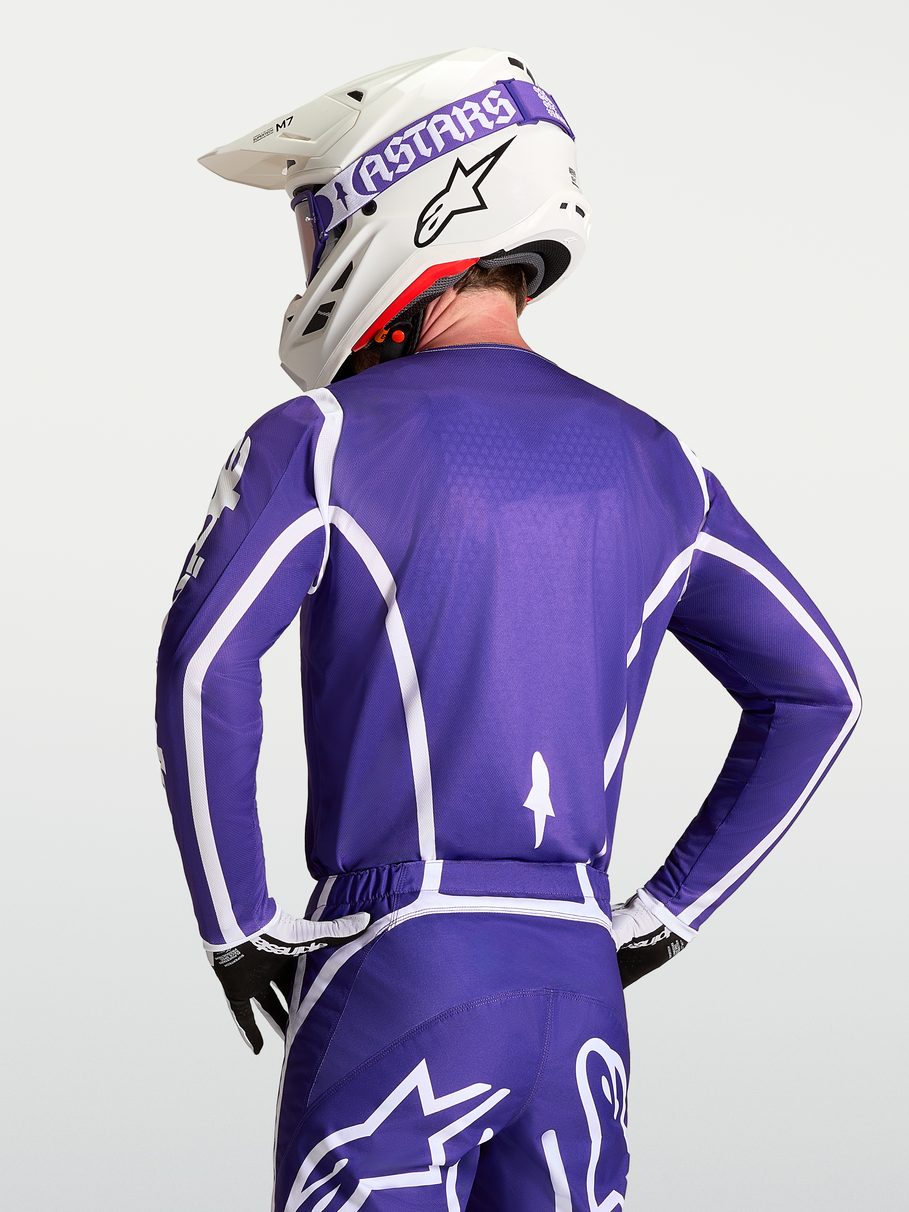 Alpinestars Koszulka Fluid Apex V2, męska koszulka motocrossowa, fioletowa i biała, oddychająca konstrukcja z poliestru, widok od tyłu na zawodnika w kasku i rękawicach.