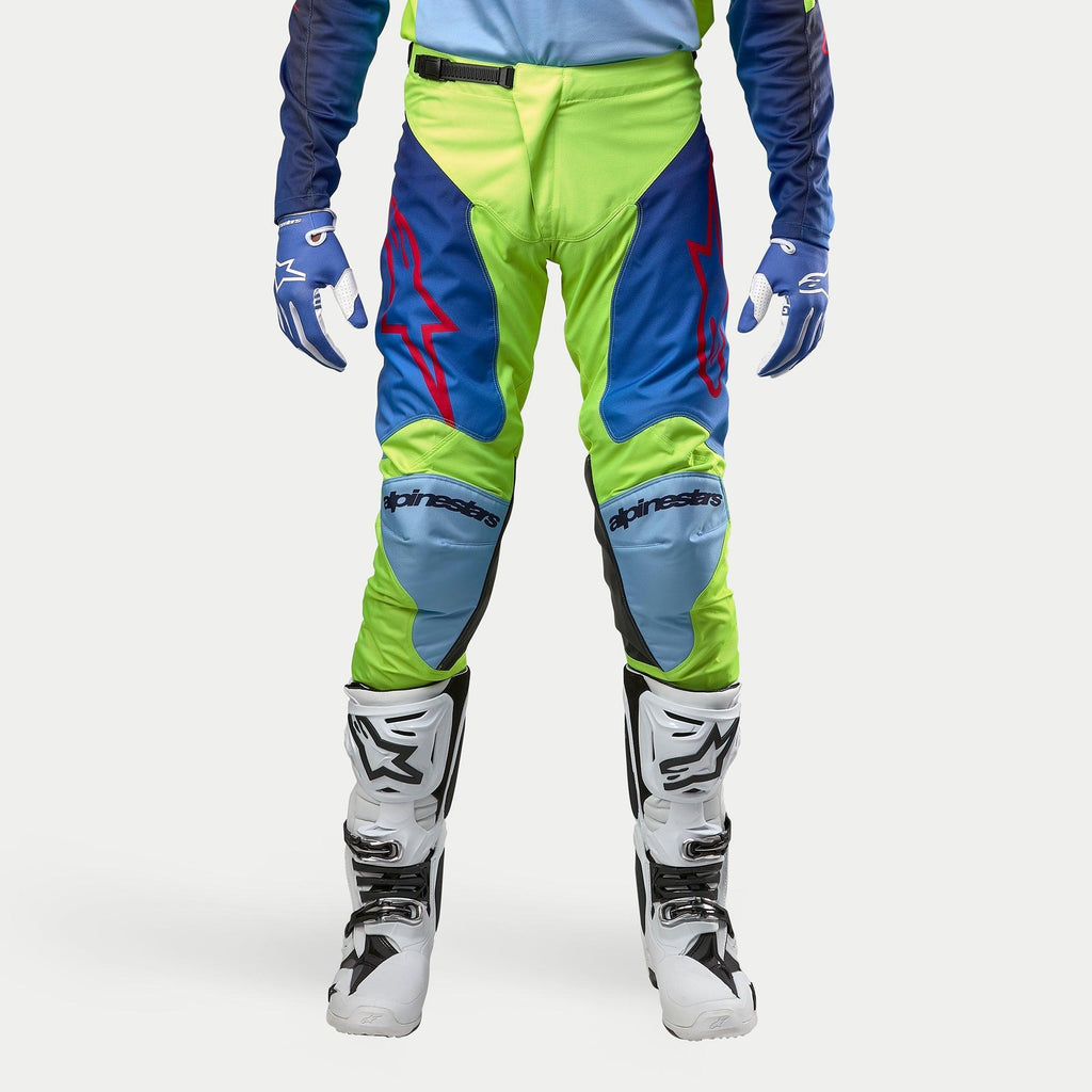 Alpinestars Racer Hoen Pants 2024, spodnie motocrossowe, żółty fluo i granatowy, noszone z biało-czarnymi butami motocrossowymi i niebieskimi rękawicami, z wyraźnymi czerwonymi logo, jasnoniebieskimi panelami na kolanach i ergonomicznym sportowym designem do jazdy w terenie