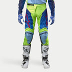 Alpinestars Racer Hoen Pants 2024, spodnie motocrossowe, żółty fluo i granatowy, noszone z biało-czarnymi butami motocrossowymi i niebieskimi rękawicami, z wyraźnymi czerwonymi logo, jasnoniebieskimi panelami na kolanach i ergonomicznym sportowym designem do jazdy w terenie