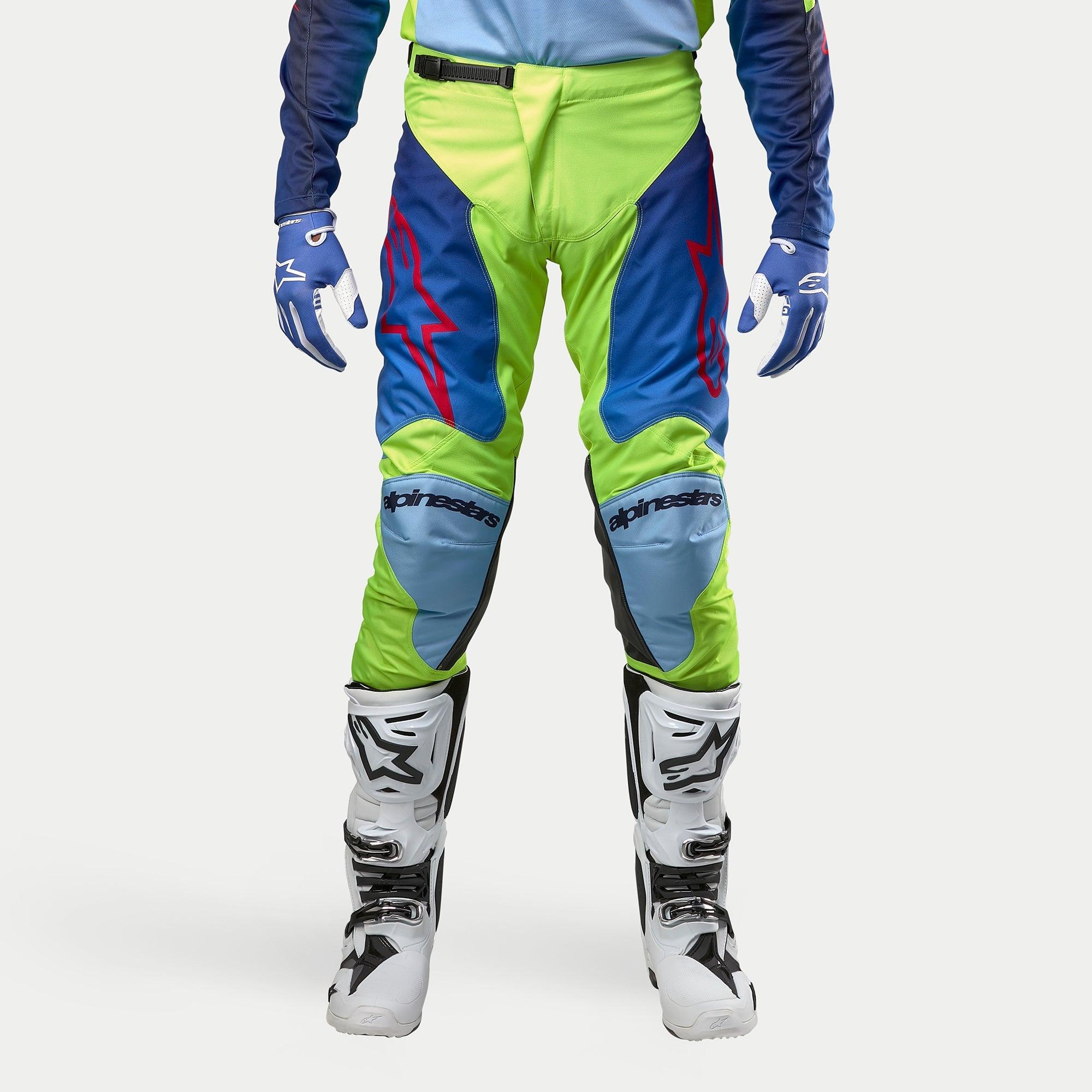 Alpinestars Racer Hoen Pants 2024, spodnie motocrossowe, żółty fluo i granatowy, noszone z biało-czarnymi butami motocrossowymi i niebieskimi rękawicami, z wyraźnymi czerwonymi logo, jasnoniebieskimi panelami na kolanach i ergonomicznym sportowym designem do jazdy w terenie