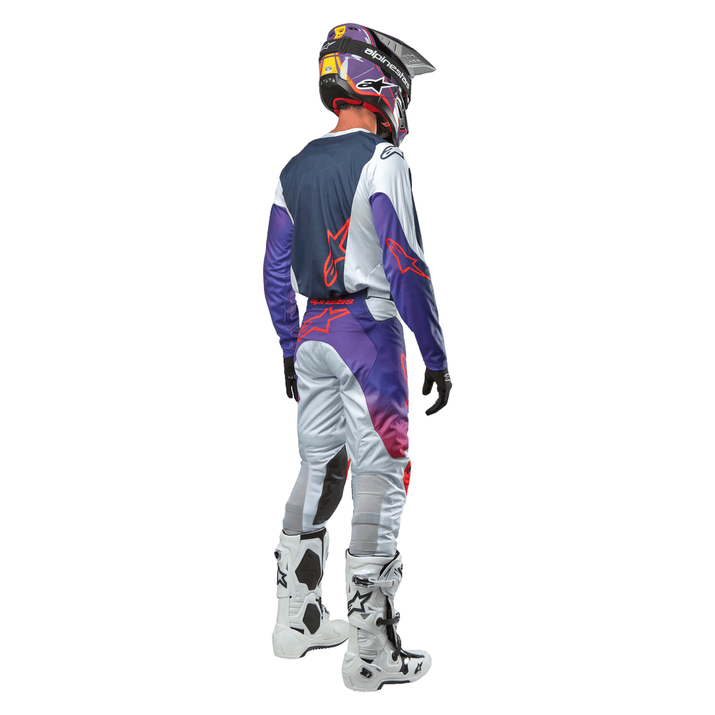 Zawodnik motocrossowy widziany od tyłu, ubrany w koszulkę Alpinestars 2024 Racer Hoen Jersey w kolorze Light Gray Hot Orange Black, granatowy panel tylny z pomarańczowym logo gwiazdy, fioletowe rękawy, pasujące spodnie wyścigowe i białe buty.