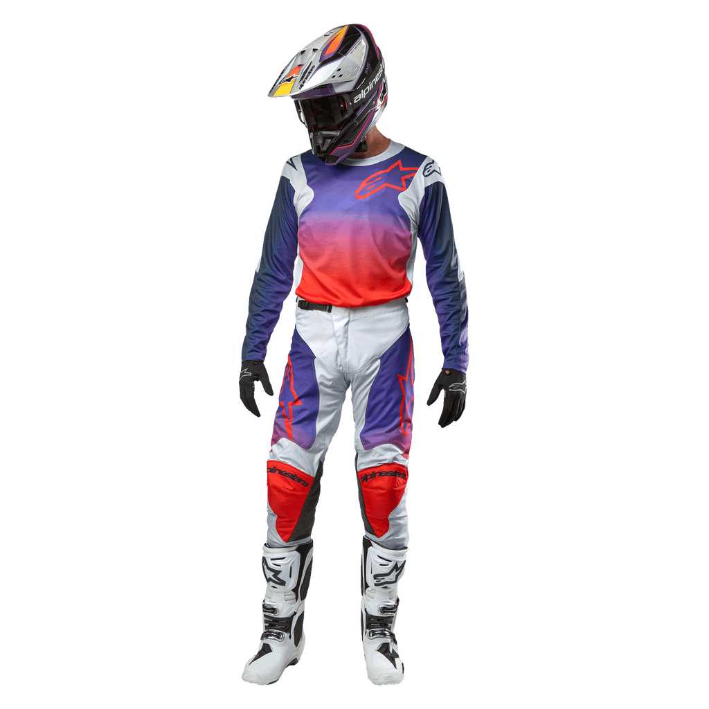 Zawodnik motocrossowy w kompletnym stroju wyścigowym, koszulka Alpinestars 2024 Racer Hoen z gradientem od fioletu do jaskrawego pomarańczu i jasnoszarymi ramionami, pasujące jasnoszare spodnie z czerwonymi i fioletowymi akcentami, dopasowany kask i białe buty, pełny zestaw do wyścigów off-road