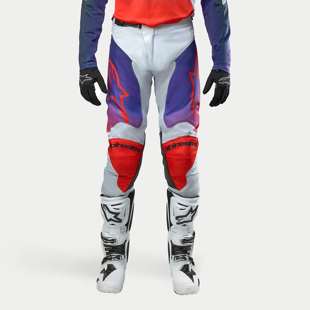 Alpinestars 2024 Racer Hoen Pants, spodnie motocrossowe, jasnoszare z fioletowymi i czerwonymi akcentami, czerwone logo na udach i ochraniacze kolan, białe buty motocyklowe, czarne rękawice, nowoczesny design sportowy